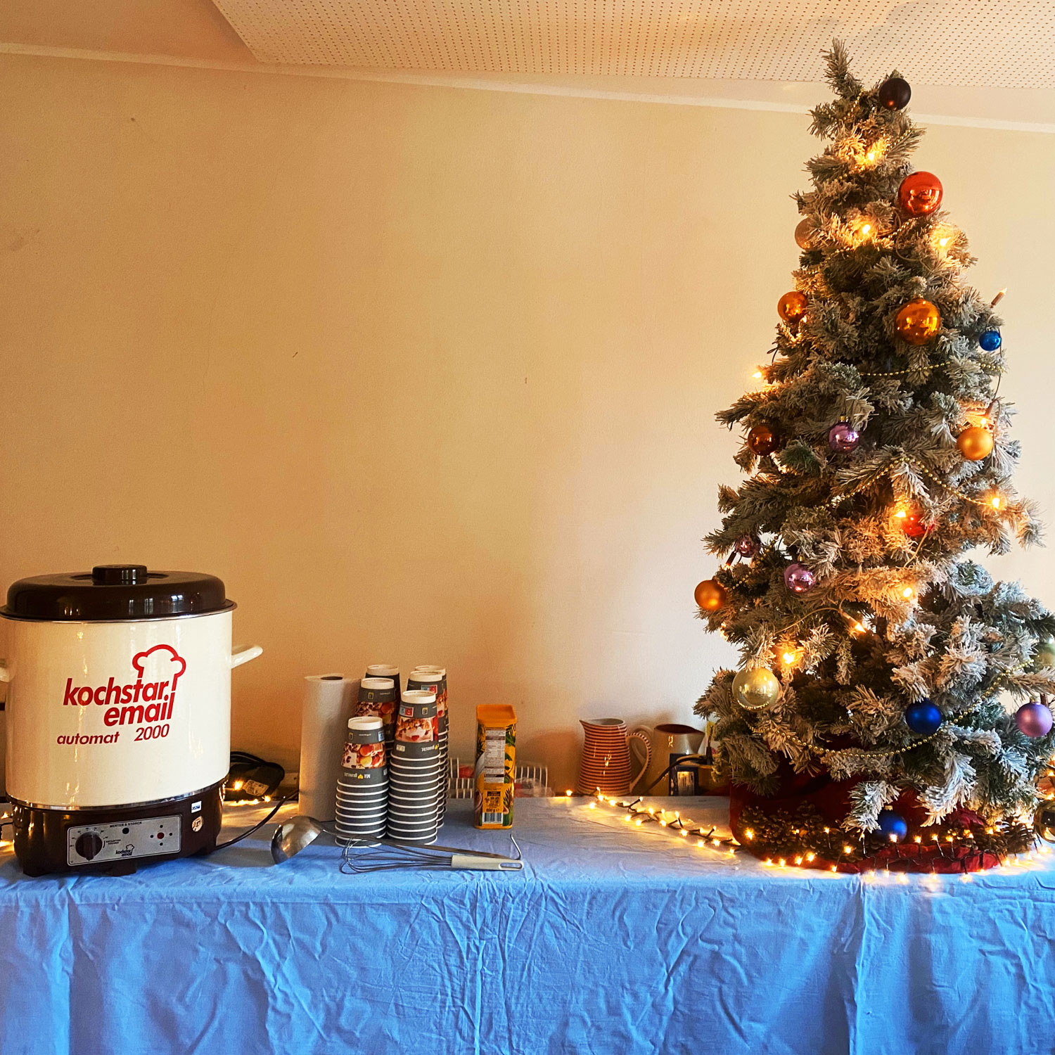 Ein Topf für warme Getränke, daneben Papierbecher und ein geschmückter Weihnachtsbaum